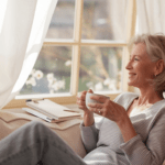 leeres Nest eine Frau sitzt beqem in einem Sessel, hat eine Tasse in der Hand. Neben sich liegt ein Notizbuch auf einem kleinen Tisch.