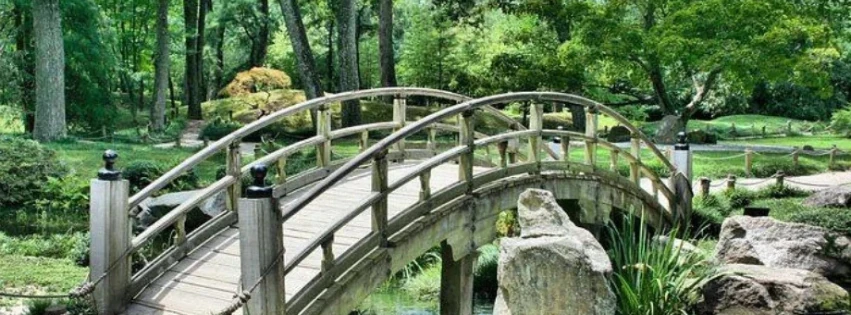 BrückeNeu Selbstverwirklichung Frauen 45 Plus Eine Brücke über einen Wasserlauf anscheinend in einer Parklandschaft mit Wegen, Wiesen, Bäumen und Felssteinen am Wasserlauf
